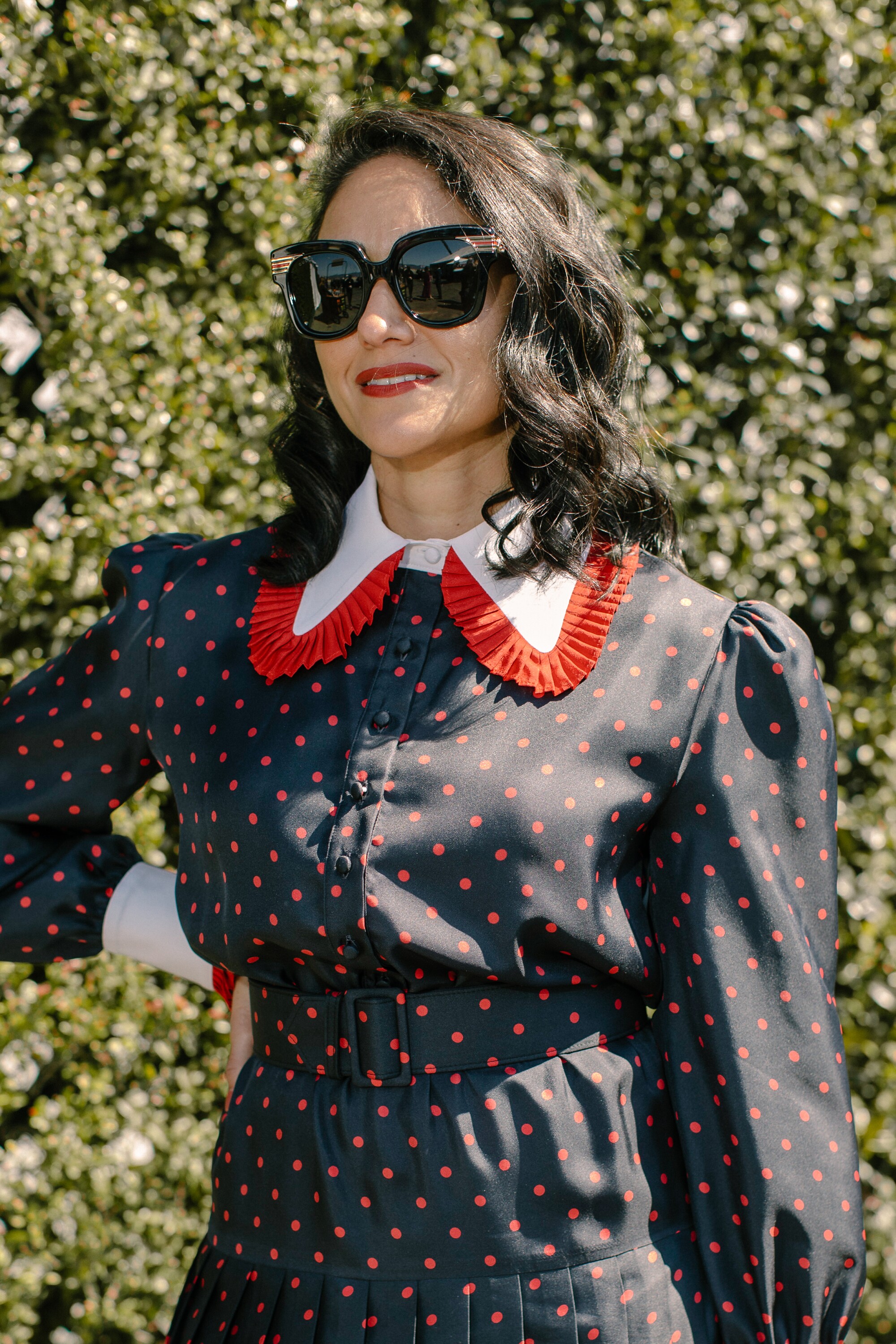 A woman in sunglasses and a dark dress with red dots and a big red and white collar.