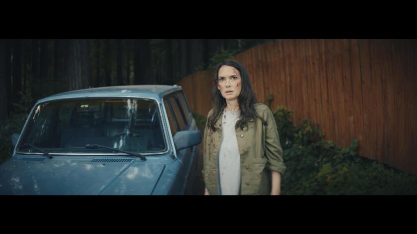 A woman with a little blood on her face stands next to a blue car 