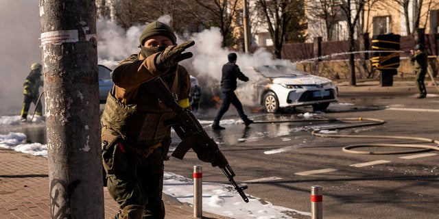 Ukrainian soldiers take positions outside a military facility as two cars burn, in a street in Kyiv, Ukraine, Saturday, Feb. 26, 2022. Russian troops stormed toward Ukraine's capital Saturday, and street fighting broke out as city officials urged residents to take shelter. 