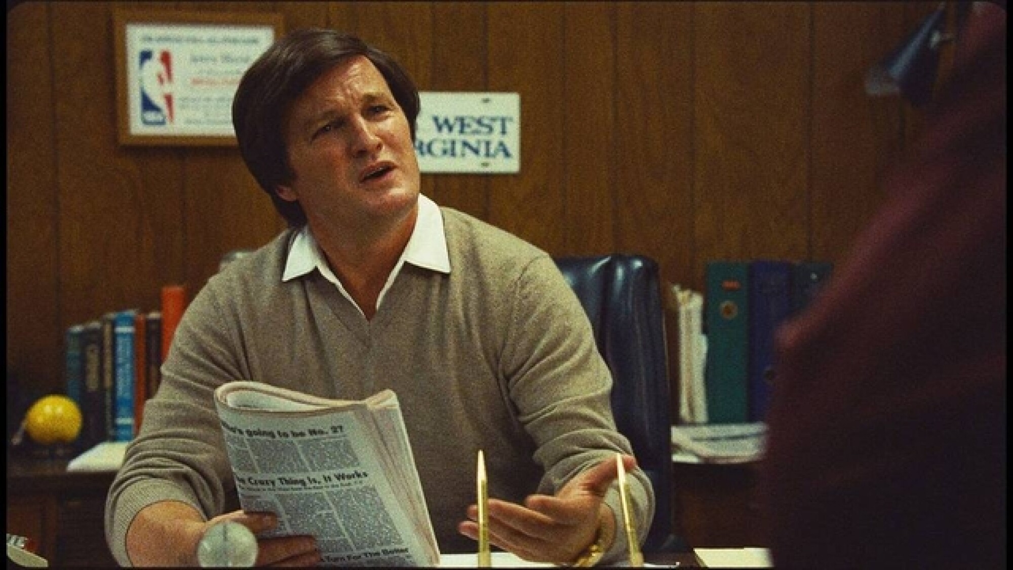 A man in a brown sweater sitting at a desk, holding a newspaper looking upset