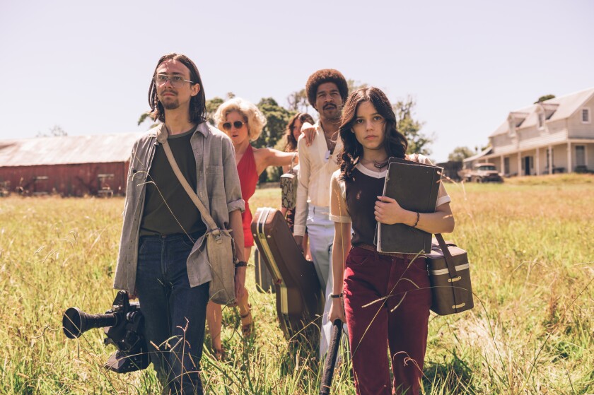 Five people walk across a field with a barn and a farmhouse in the background.