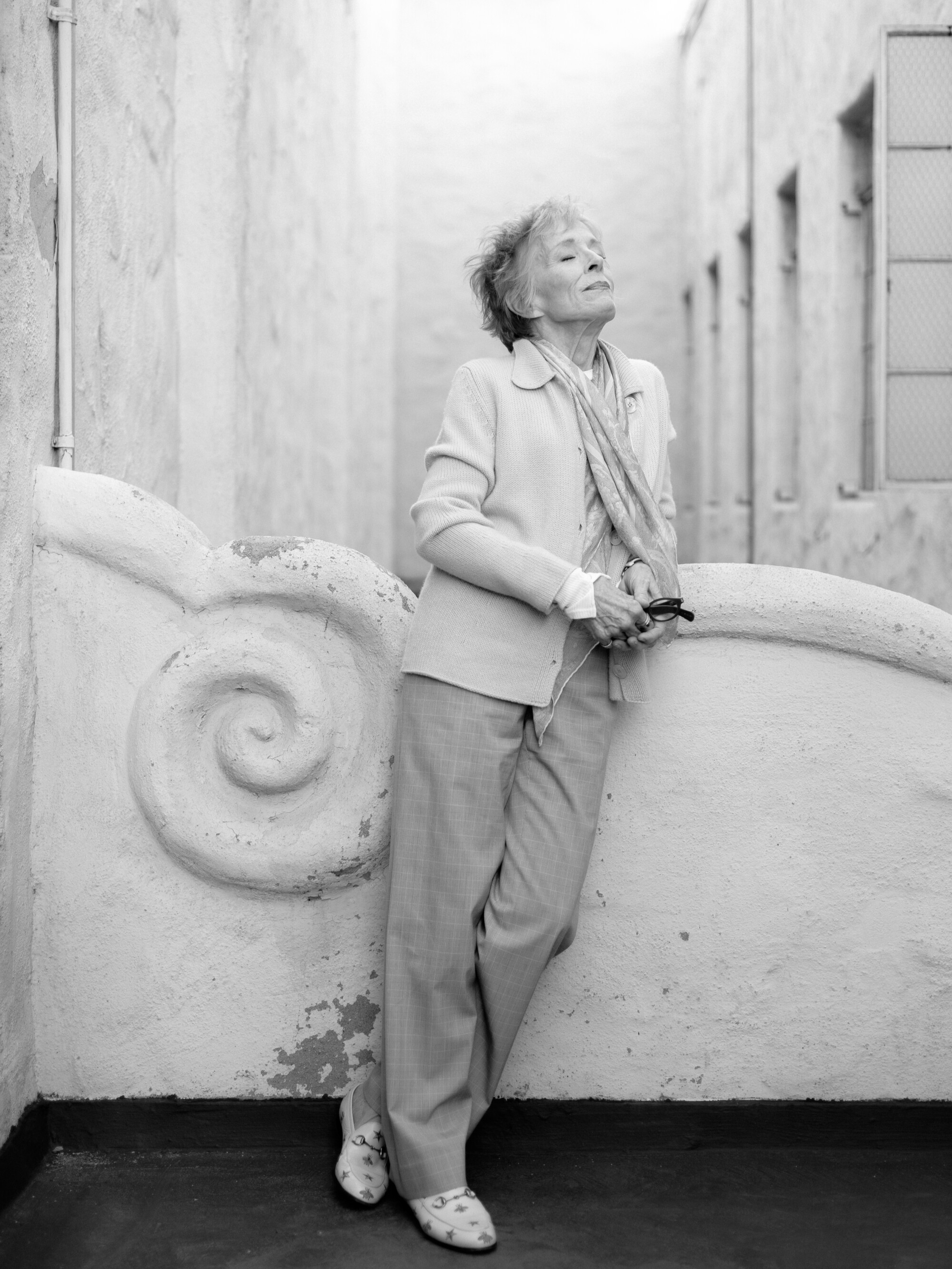 A black-and-white portrait of a standing woman leaning against an ornate wall.