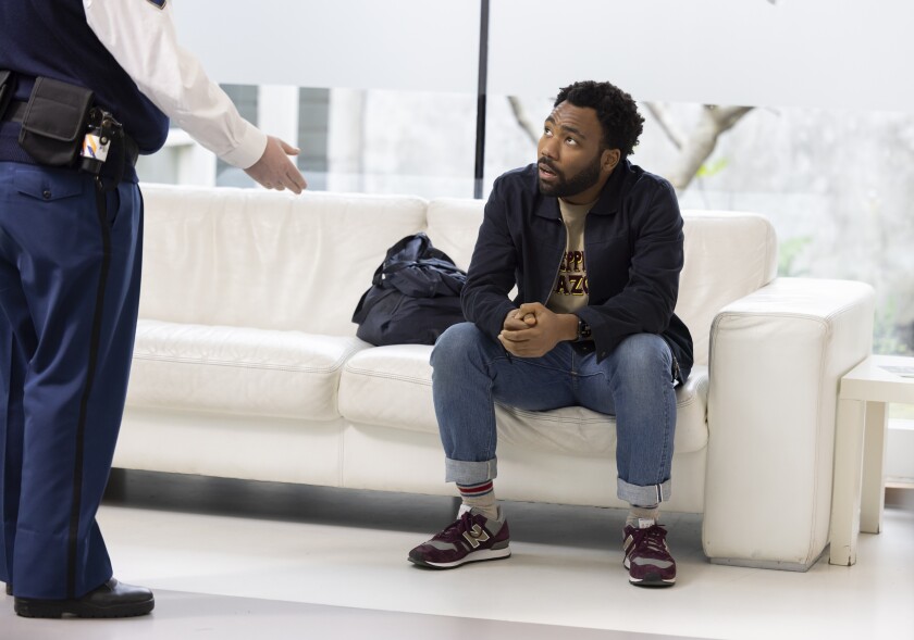 A man sitting on a white sofa as another man gestures at him.