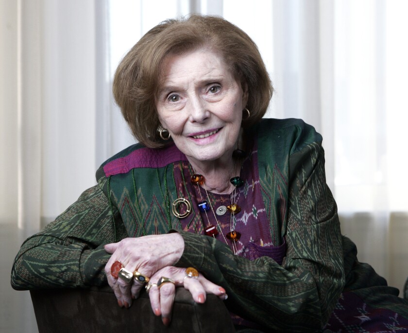 An older woman in a green suit leans on one arm on the back of a chair