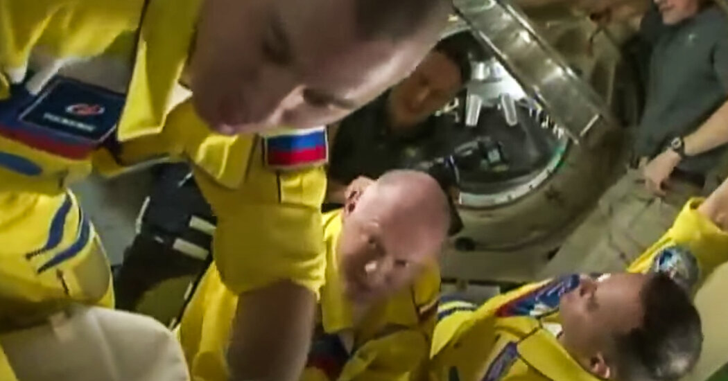 Russia’s astronauts enter the space station in yellow and blue flight suits.