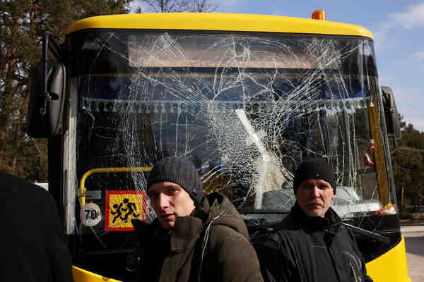 Two men stand in front of a bush with a spiderwebbed windshield.