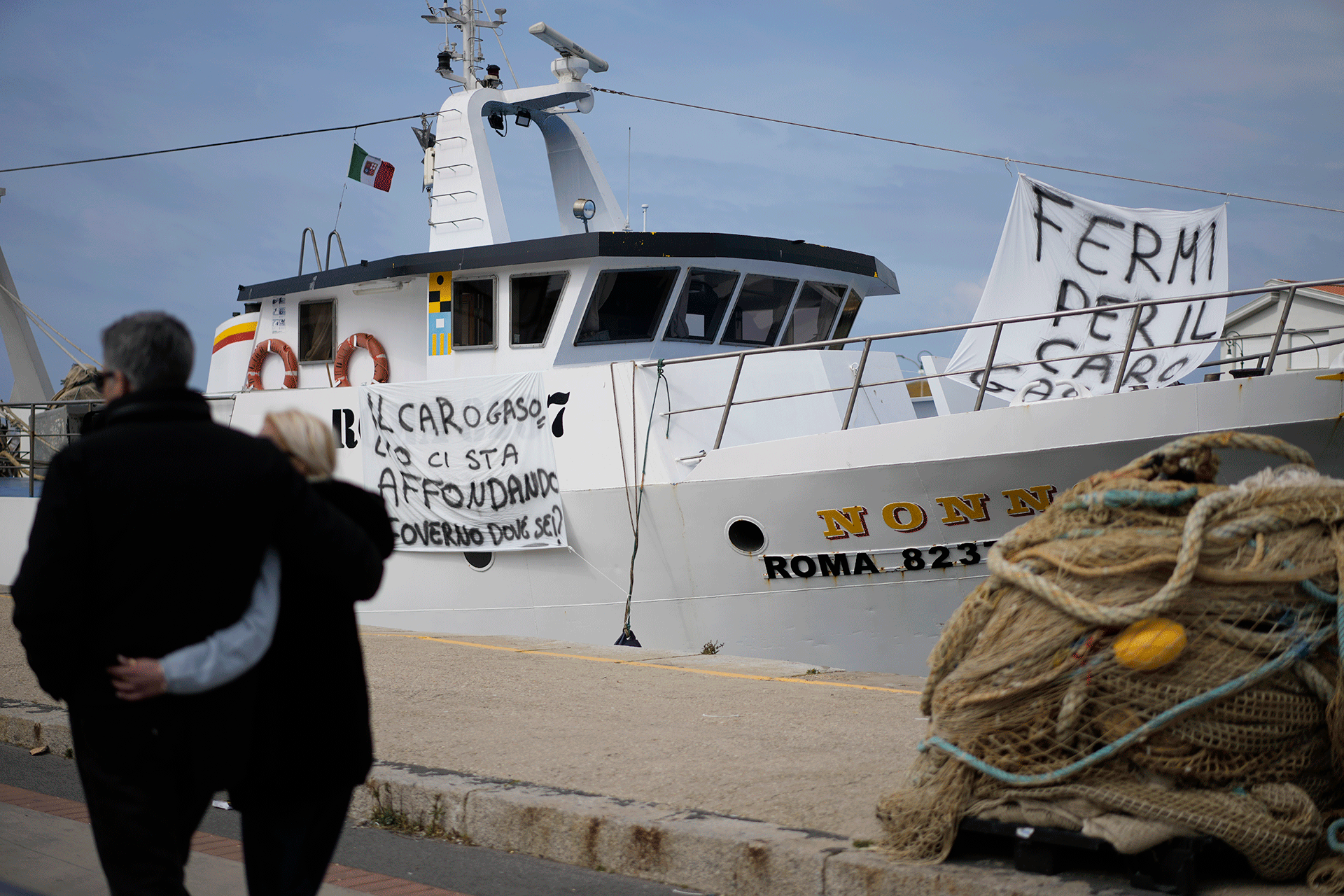 People walk past a fishing boat with banners against the gasoline price increase in the Roman port of Fiumicino, Friday, March 11, 2022.