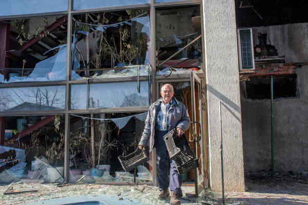 A man holds crates in front of a damaged, modern-looking building.