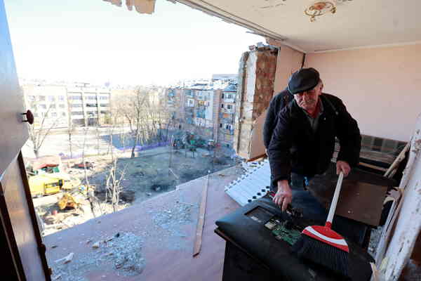 A man cleans a computer monitor in a high-rise apartment. One wall of the apartment is missing and open to the elements.
