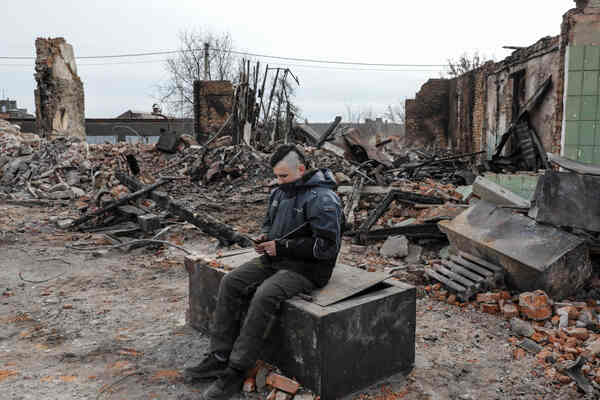 A man sits between broken houses.