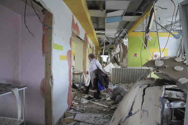 A white-coated person walks through a pastel-colored hallway. Walls, doors and ceilings are broken; wires hang to the floor.