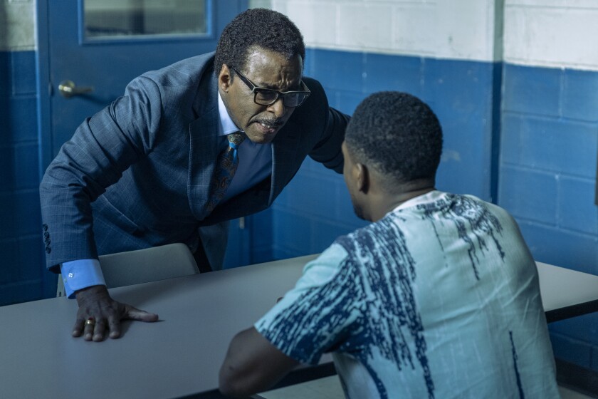 A man in a suit leans on a table as he interrogates an imprisoned man.