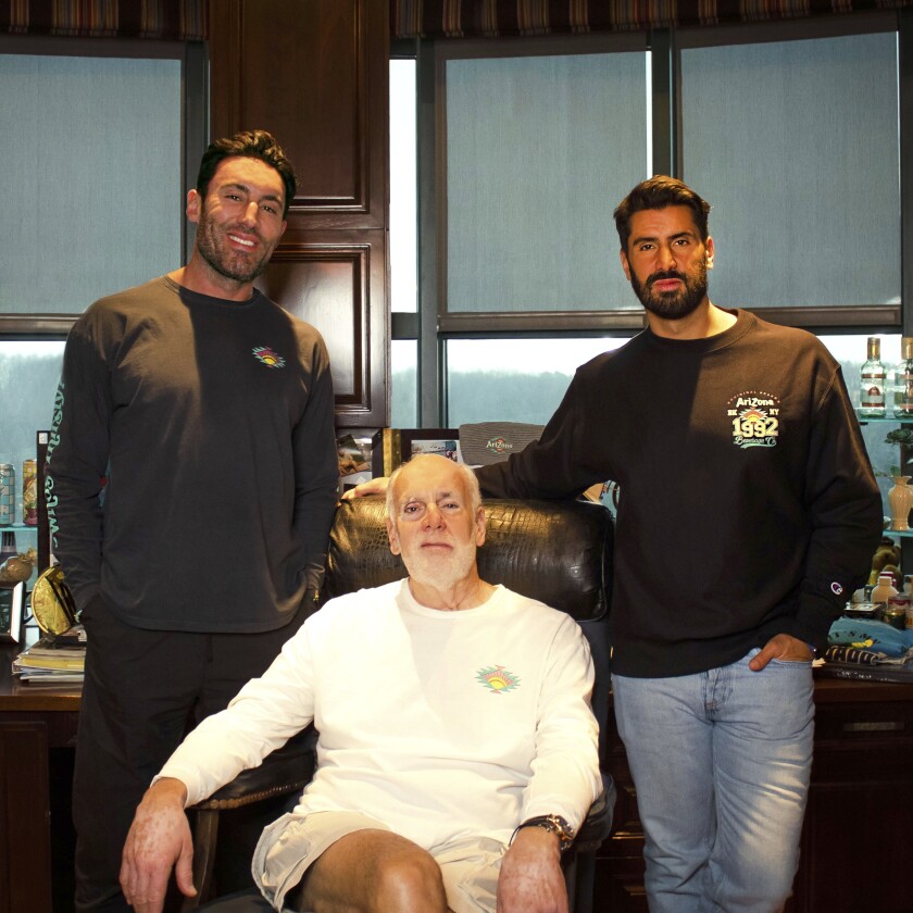 An older man with a beard, seated, is flanked by two younger men with dark hair who are standing.