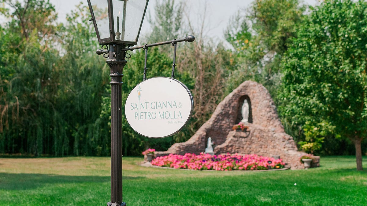 Sign reading "Saint Gianna and Pietro Molla Maternity Home" in front of grotto