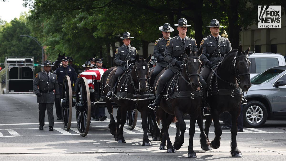 Four horses with mounted police trot down the street with a hearse carriage in tow