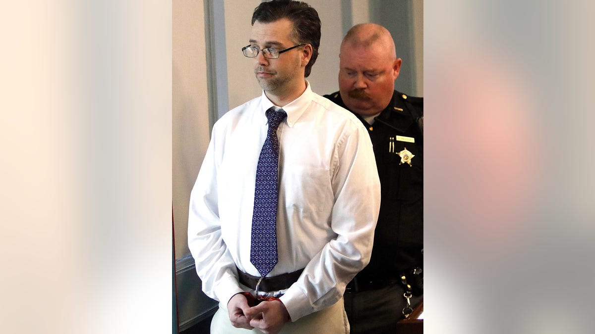 Shawn Grate in a white shirt and blue tie handcuffed in front of a cop.