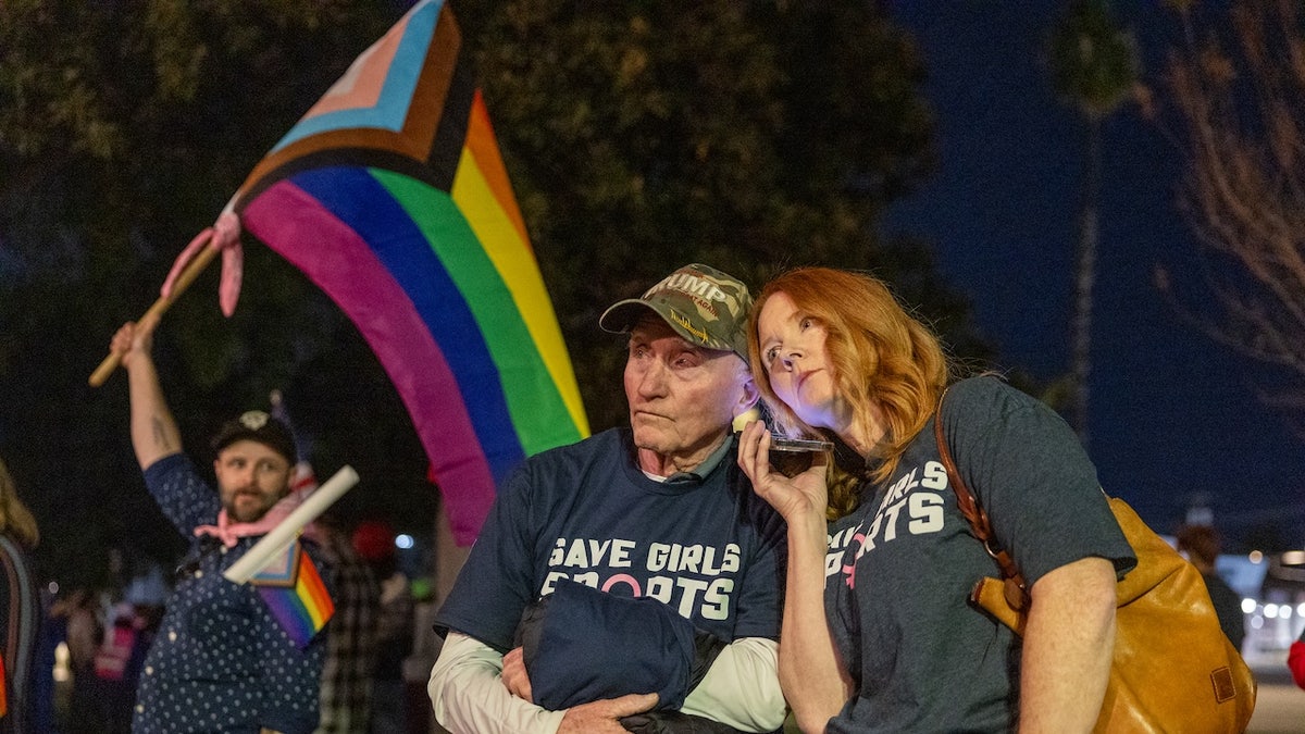 Transgender athlete supporter Kyle Harp, left, of Riverside holds the progress pride flag as "Save Girls Sports" supporters Lori Lopez and her dad Pete Pickering, both of Riverside, listen to the debate as they join the overflow crowd converging outside the Riverside Unified School District meeting Thursday night to debate the rights of transgender athletes to compete in high school sports, Thursday, Dec. 19, 2024. 